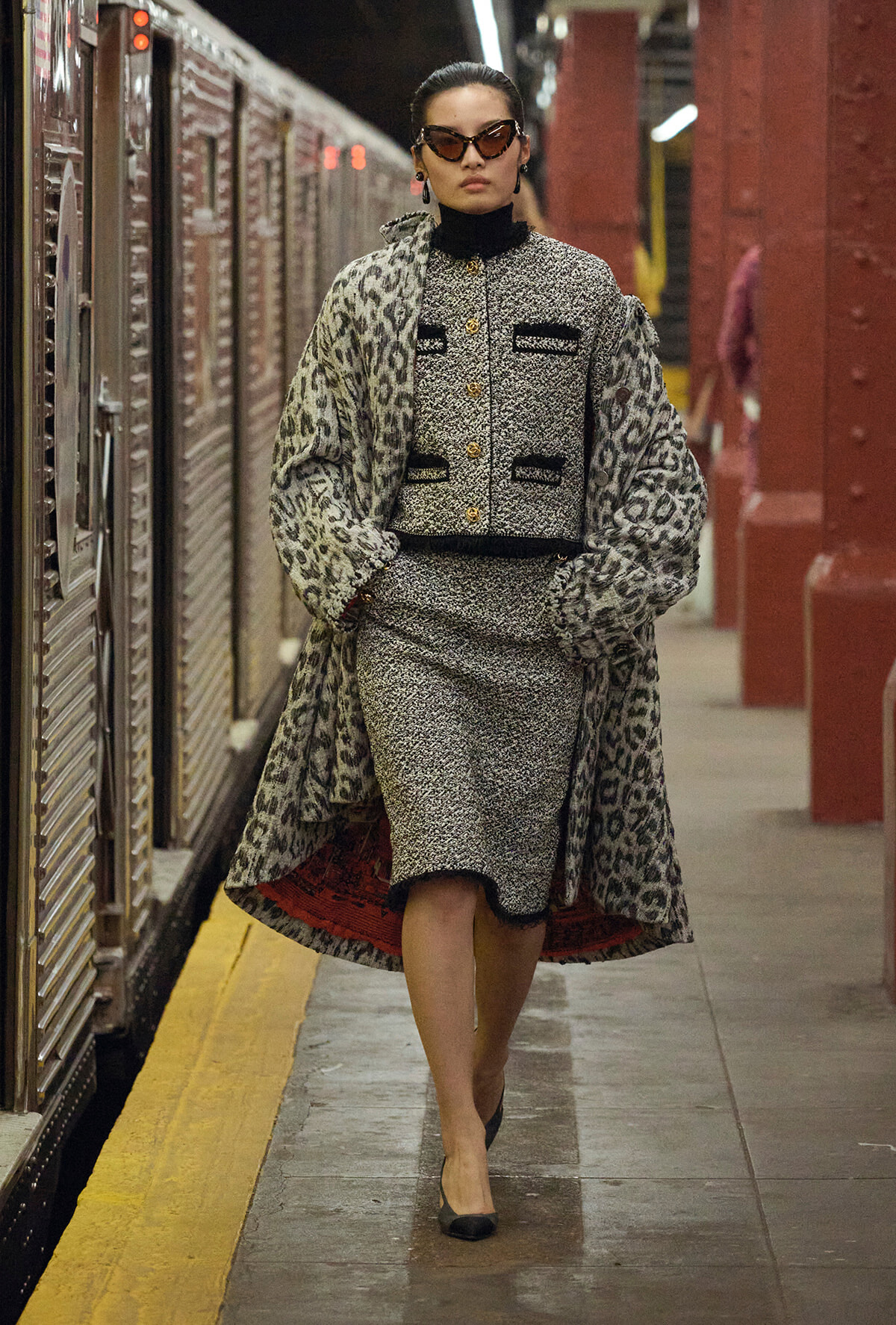 Fashionable woman in a leopard-print coat and matching outfit walks along a subway platform with a train to the side and yellow safety line nearby.