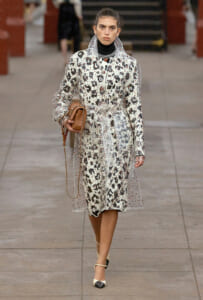 Female model walks a fashion runway wearing a white sheer floral-printed trench coat over a black turtleneck, holding a tan leather quilted handbag with a gold chain.