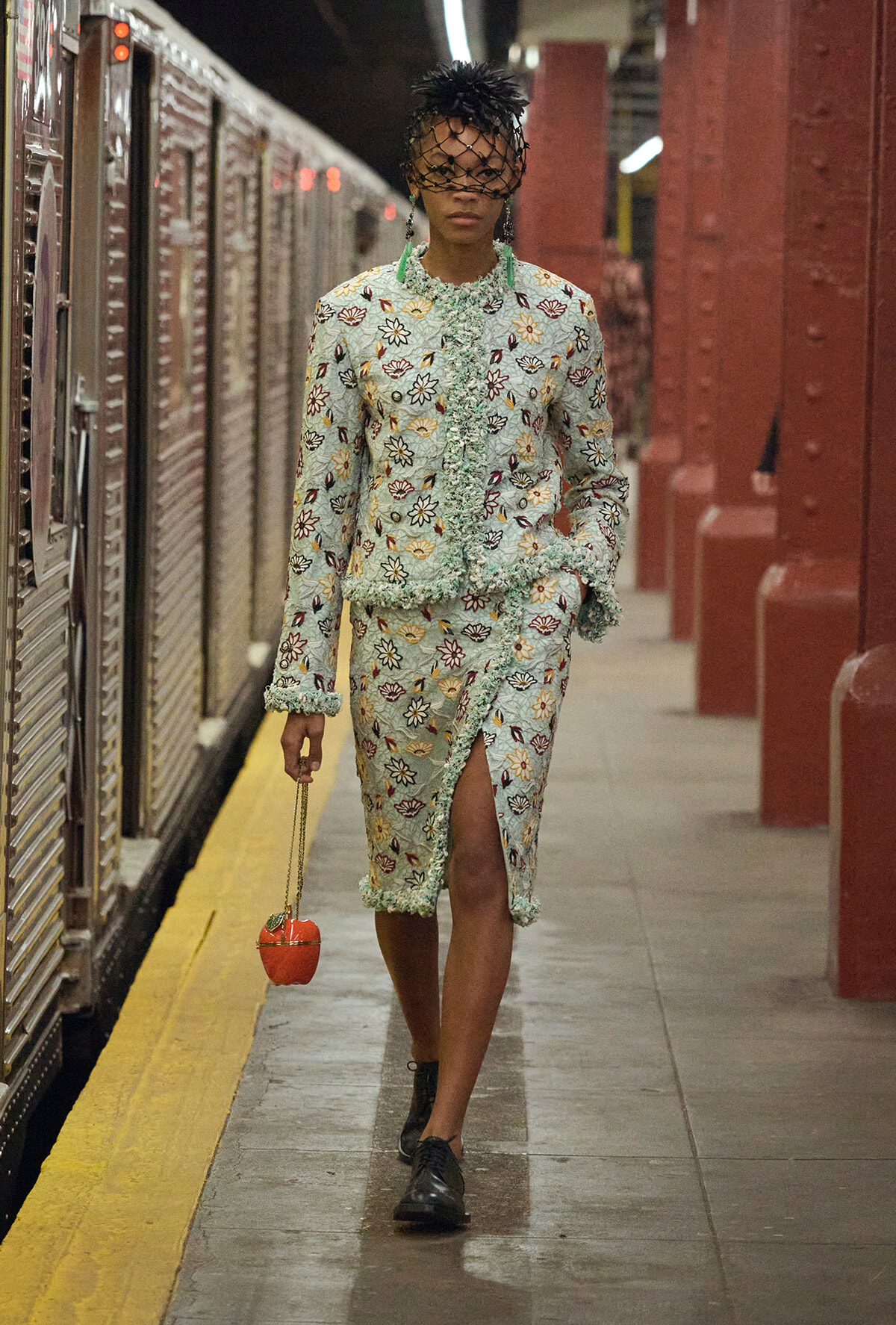 Model in a mint floral suit with fringe, wearing a net veil, on a subway platform with an orange handbag.