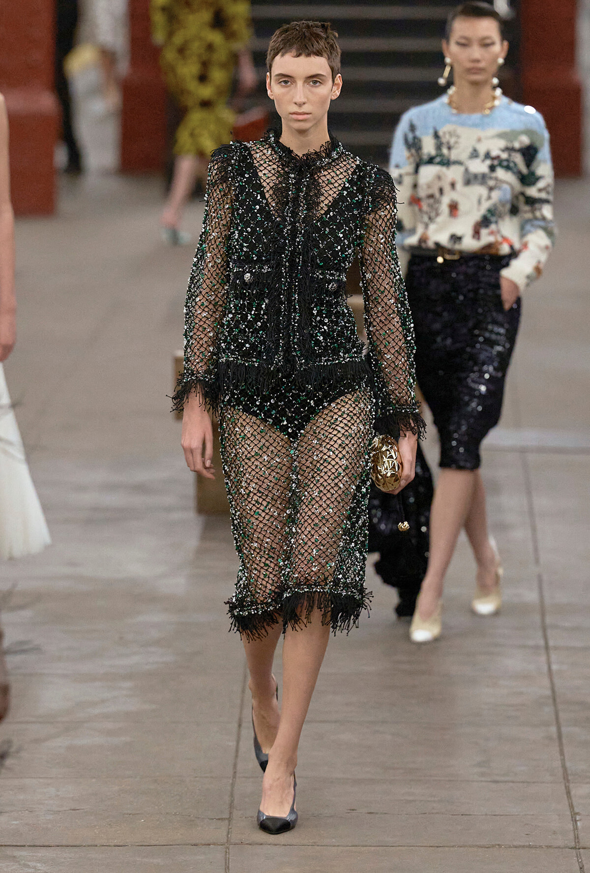 Model walking a runway in a sheer black beaded dress with fringe, holding a gold clutch with a geometric pattern
