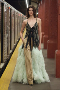 Model wearing a geometric patterned dress with a long mint feather train, standing on a subway platform beside a train car.