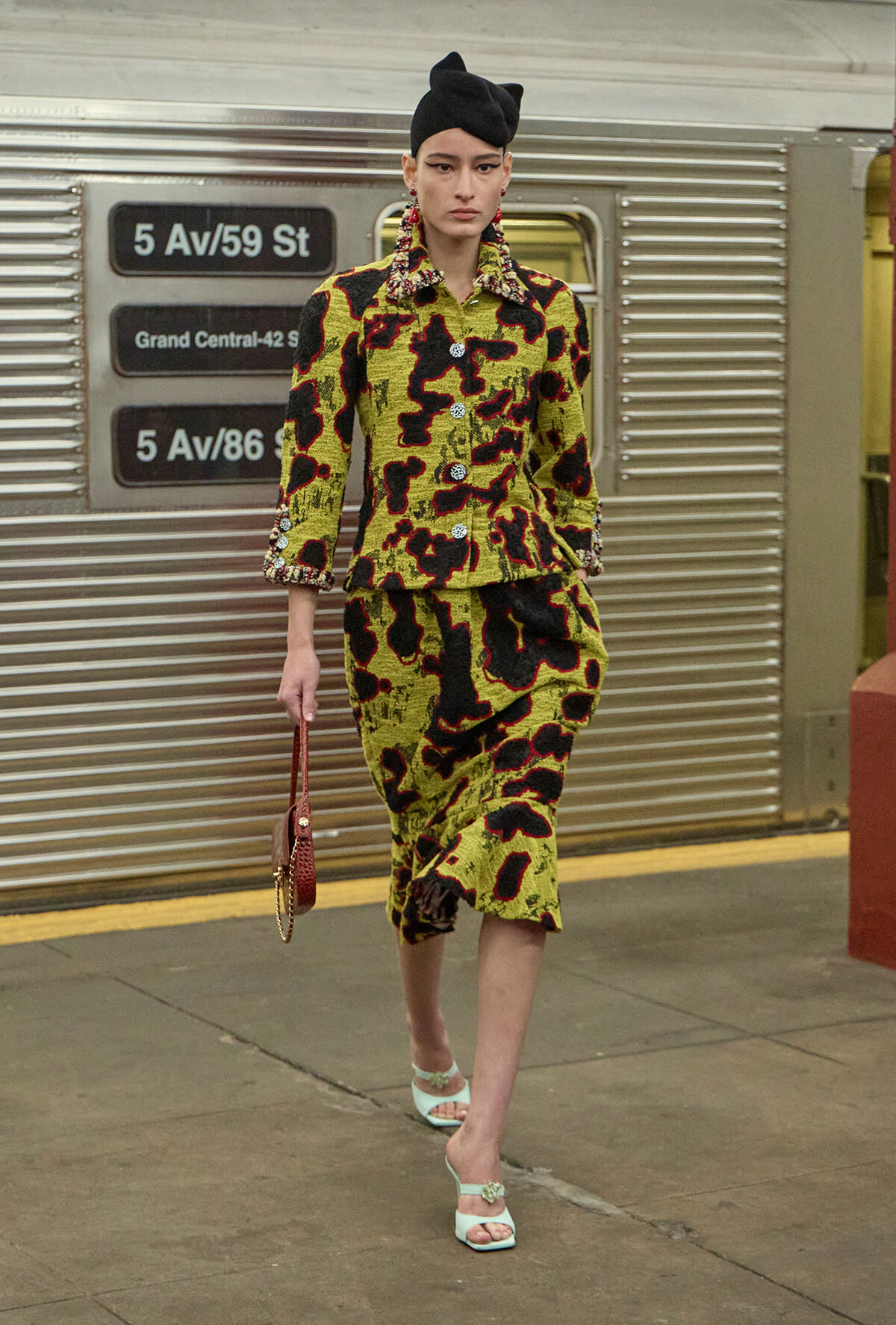 Model wearing a yellow-green, black abstract-print suit with a matching skirt on a subway platform, accessorized with a black turban-style hat, statement earrings, a small brown purse, and mint green heeled sandals.