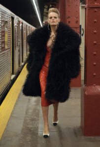 Woman in a black fur coat over a red sequin dress walking on a subway platform with white heels.",