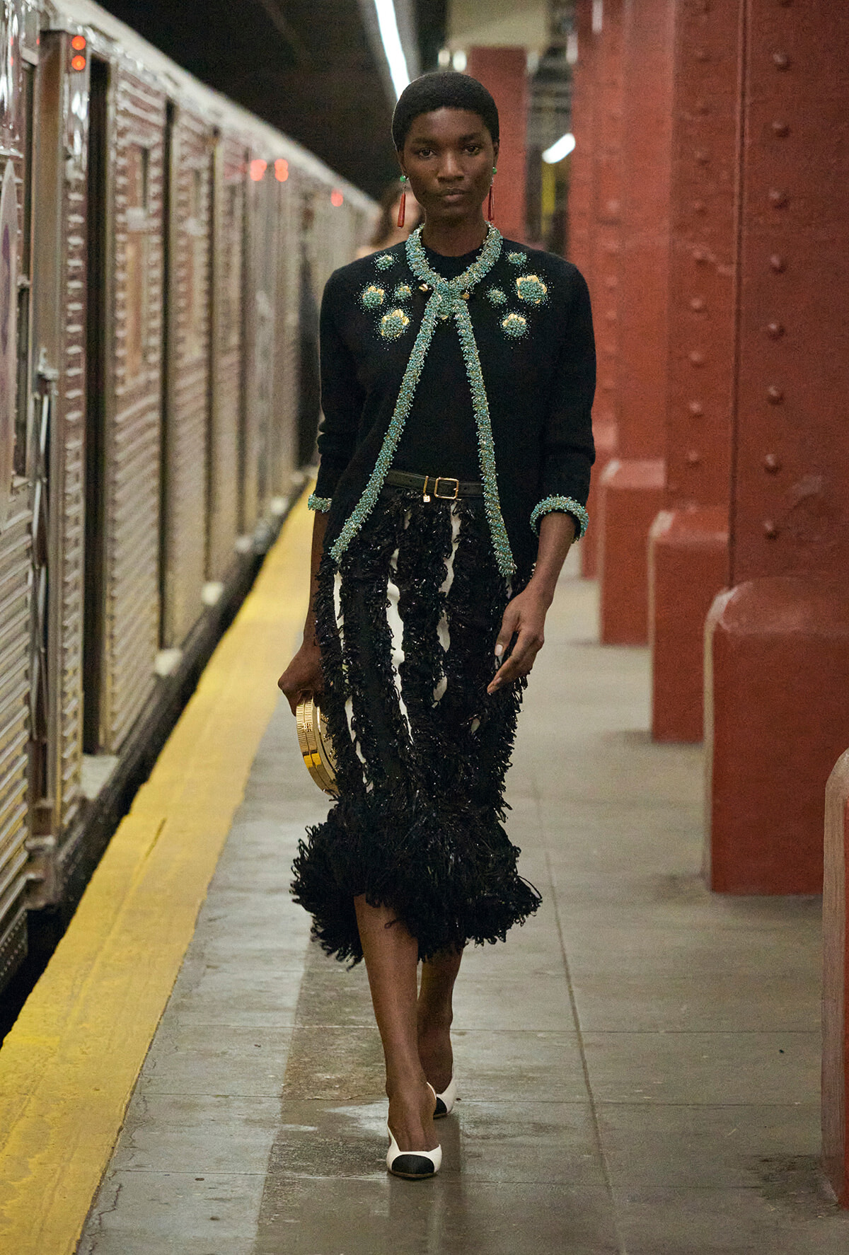 Woman in a black embellished cardigan with turquoise beading and a feathered skirt walking on a subway platform, holding a gold clutch.