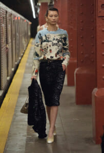 Model in a festive snowy village sweater and a black sequined midi skirt stands on a subway platform, holding a black sequined coat and beige handbag.