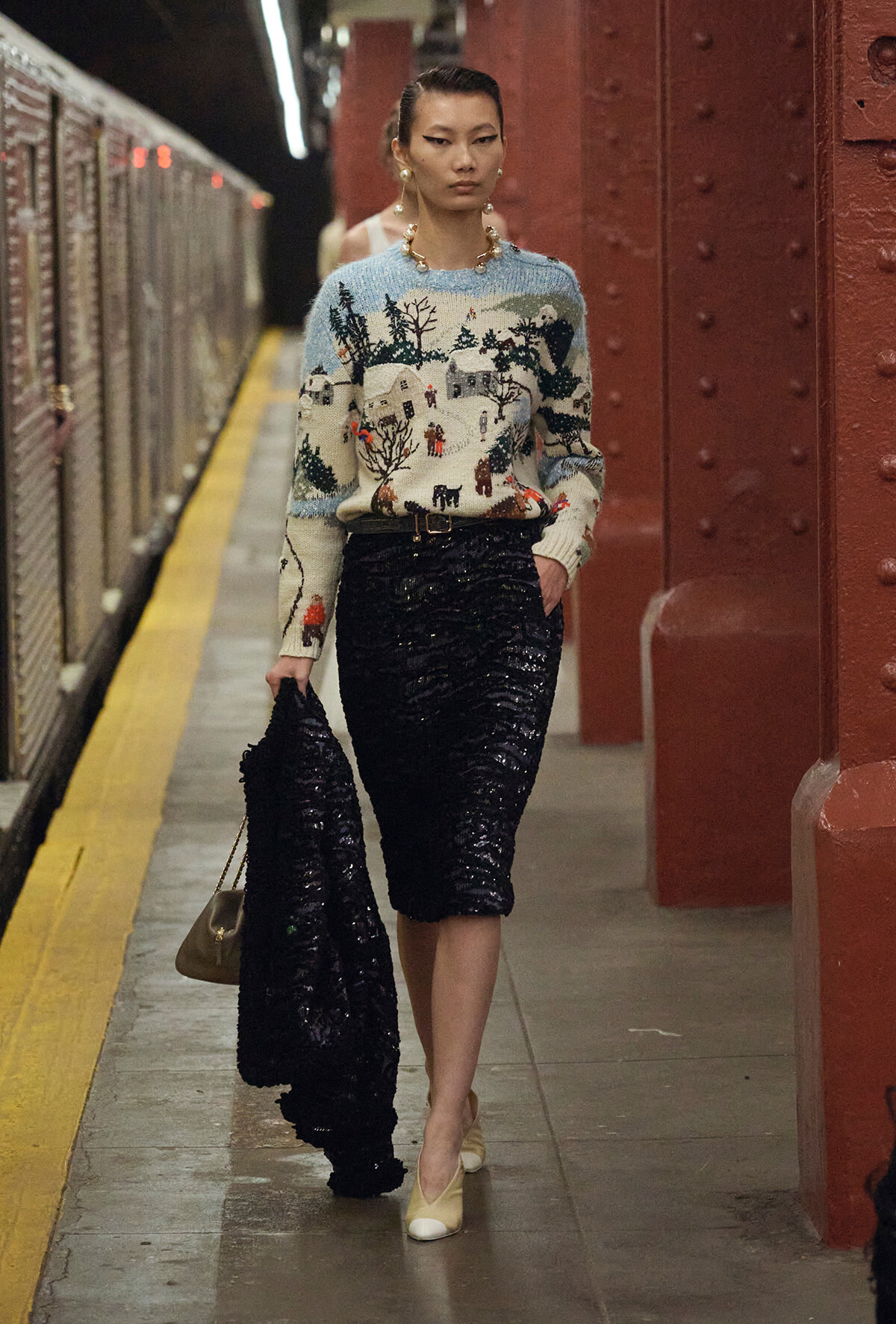Model in a festive snowy village sweater and a black sequined midi skirt stands on a subway platform, holding a black sequined coat and beige handbag.