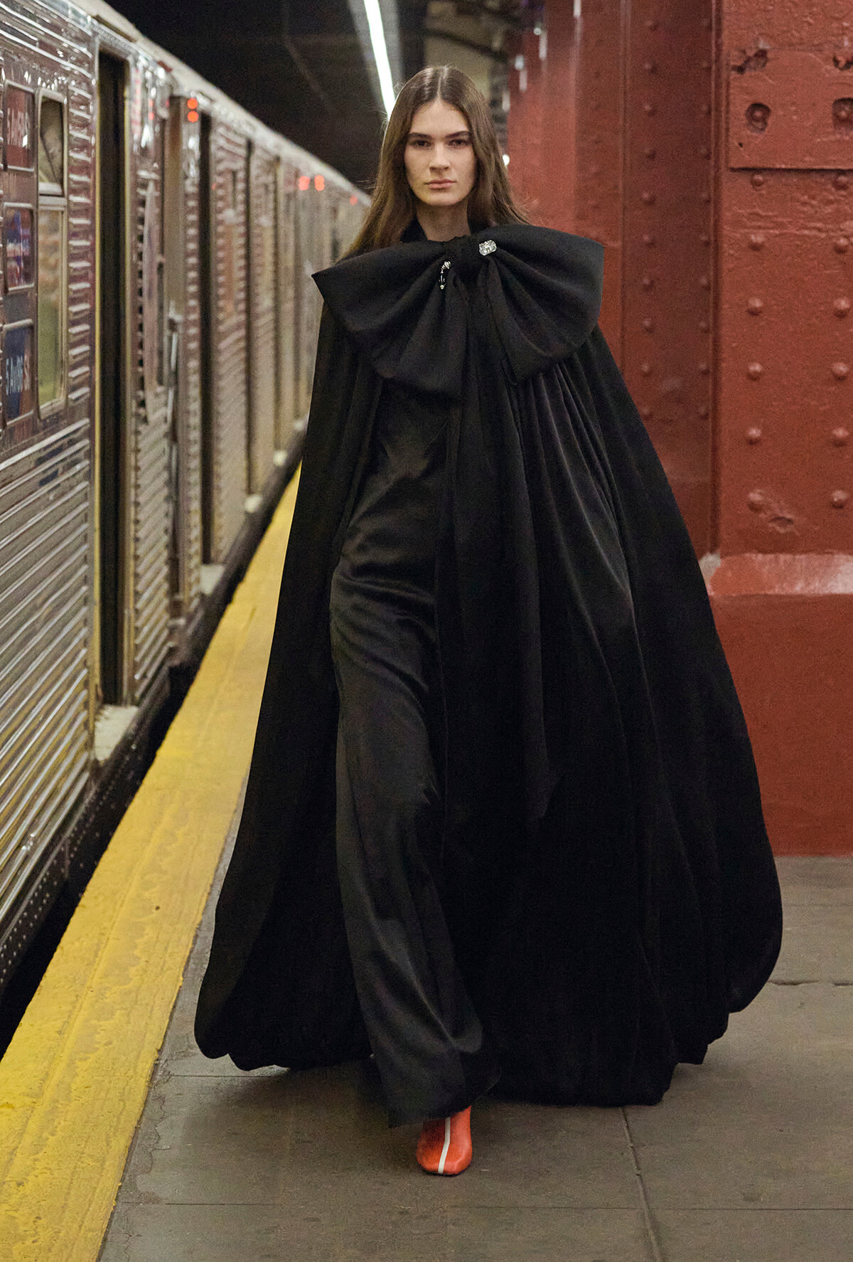 Person in a dramatic black cape with a large bow walks along a subway platform, orange-red shoes peeking out