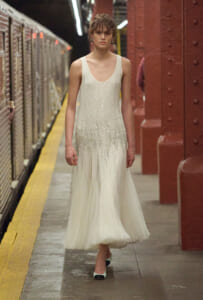 Model in a sparkling white sleeveless gown standing on a subway platform beside a yellow line.