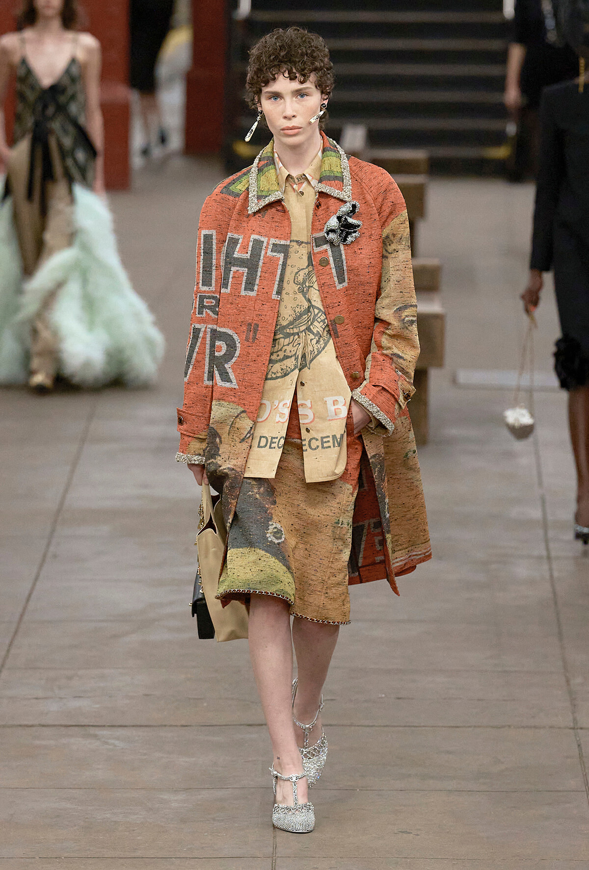 Model walking runway in a patchwork orange-brown coat over a beige printed dress and glittery heels.