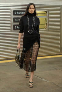 Woman in black turtleneck and lace skirt posing on a subway platform, carrying a small handbag with chain detail and layered necklaces.