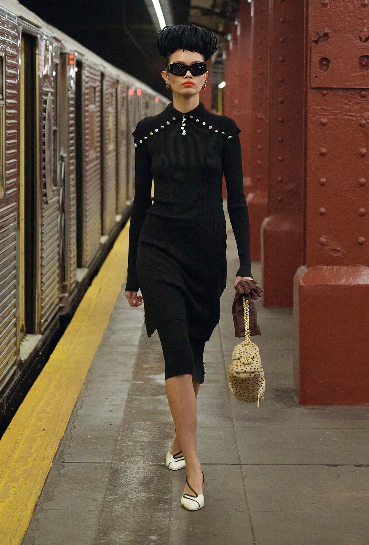 Stylish woman in a black dress with pearl accents walks along a subway platform, carrying a leopard-print bag and wearing sunglasses.