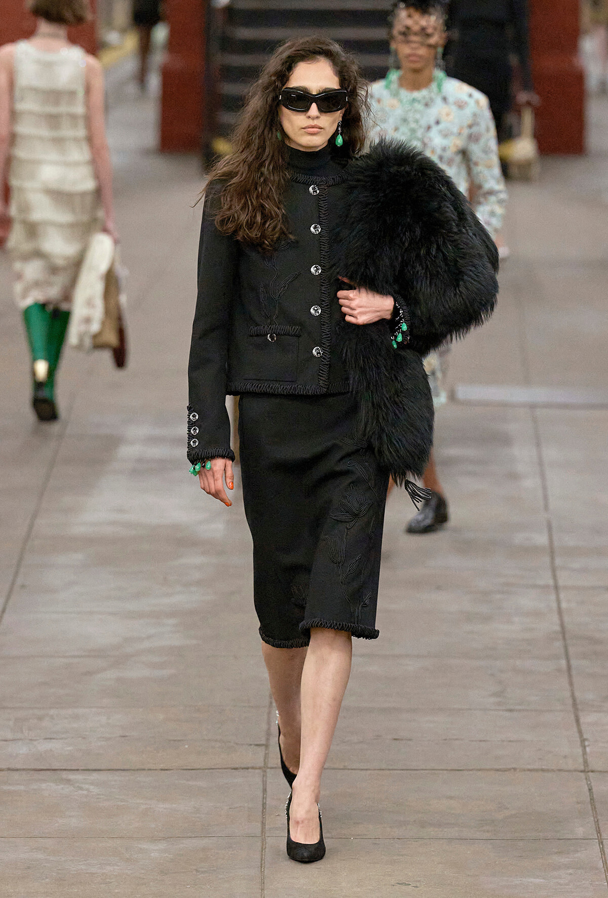 Model on a runway wearing a black tailored suit with silver buttons, holding a black fur stole and green jewelry, wearing dark sunglasses and black heels.
