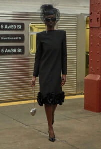 Stylish woman in a black knee-length dress with a voluminous flower hem, veil hat and sunglasses, holding a small silver heart-shaped purse at a subway station.