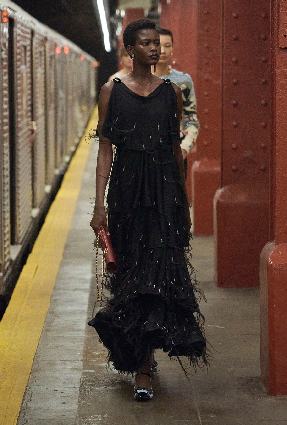 Woman in a voluminous black layered gown walking through a subway platform with red steel pillars, carrying a red clutch and chain strap bag.