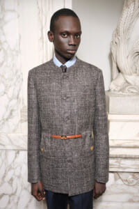 Male model in a gray tweed coat with a tan belt, blue striped shirt, and dark trousers, standing beside a marble sculpture backdrop.