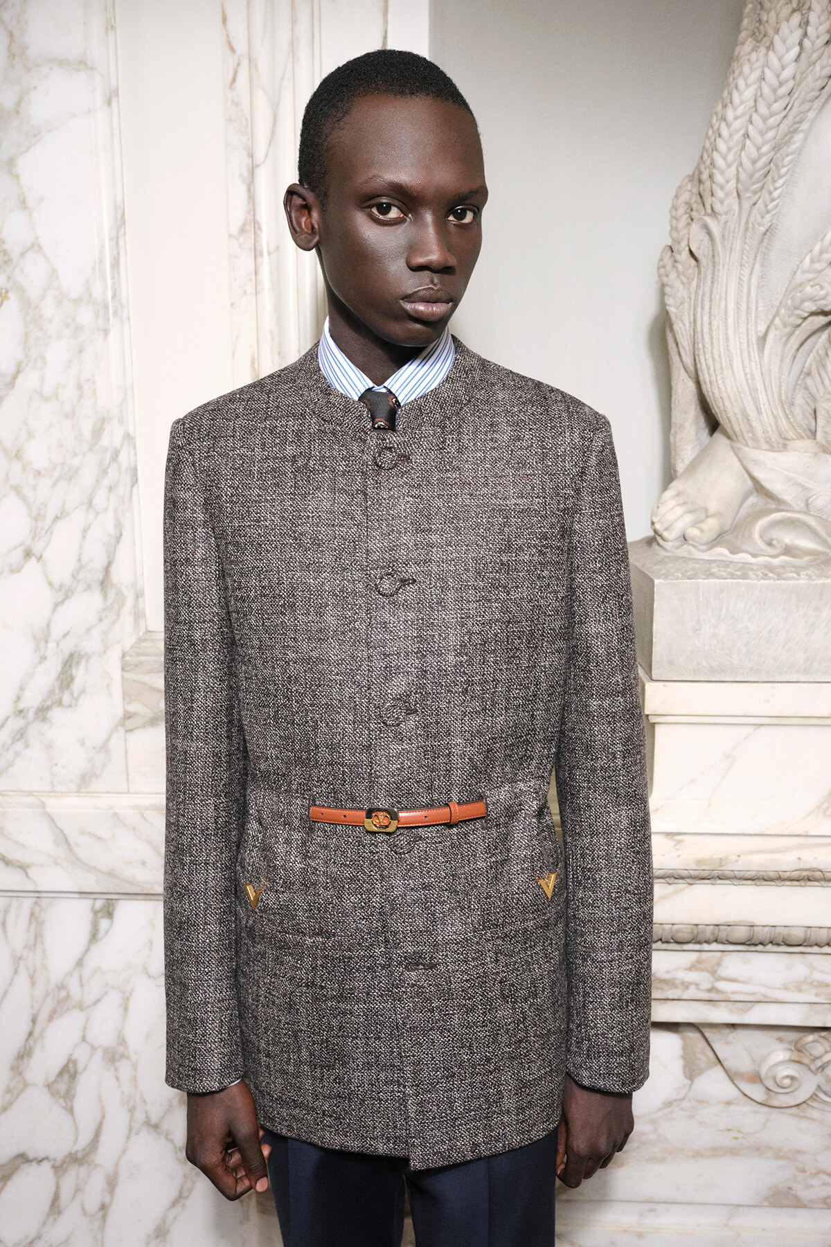 Male model in a gray tweed coat with a tan belt, blue striped shirt, and dark trousers, standing beside a marble sculpture backdrop.