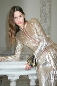Model wearing a gold‑silver striped sequin dress, leaning on a marble railing in an elegant interior.