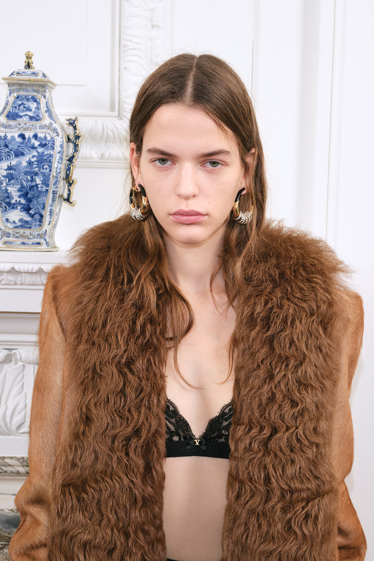 Model wearing a brown fur coat with a black lace bra visible, gold hoop earrings, standing indoors beside a blue-and-white vase on a mantelpiece.