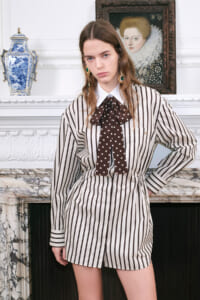 Model wearing a white shirt dress with black vertical stripes and a brown polka-dot bow, posing beside a marble fireplace in a classical room.
