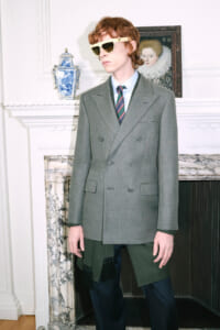 Model wearing a gray double‑breasted blazer, blue shirt, striped tie, and pale yellow sunglasses indoors by a marble fireplace.