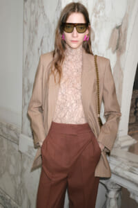 Model wearing a beige pinstripe blazer over a pink lace turtleneck, rust-colored wide trousers, pink earrings, and chunky sunglasses with a chain-strap bag.