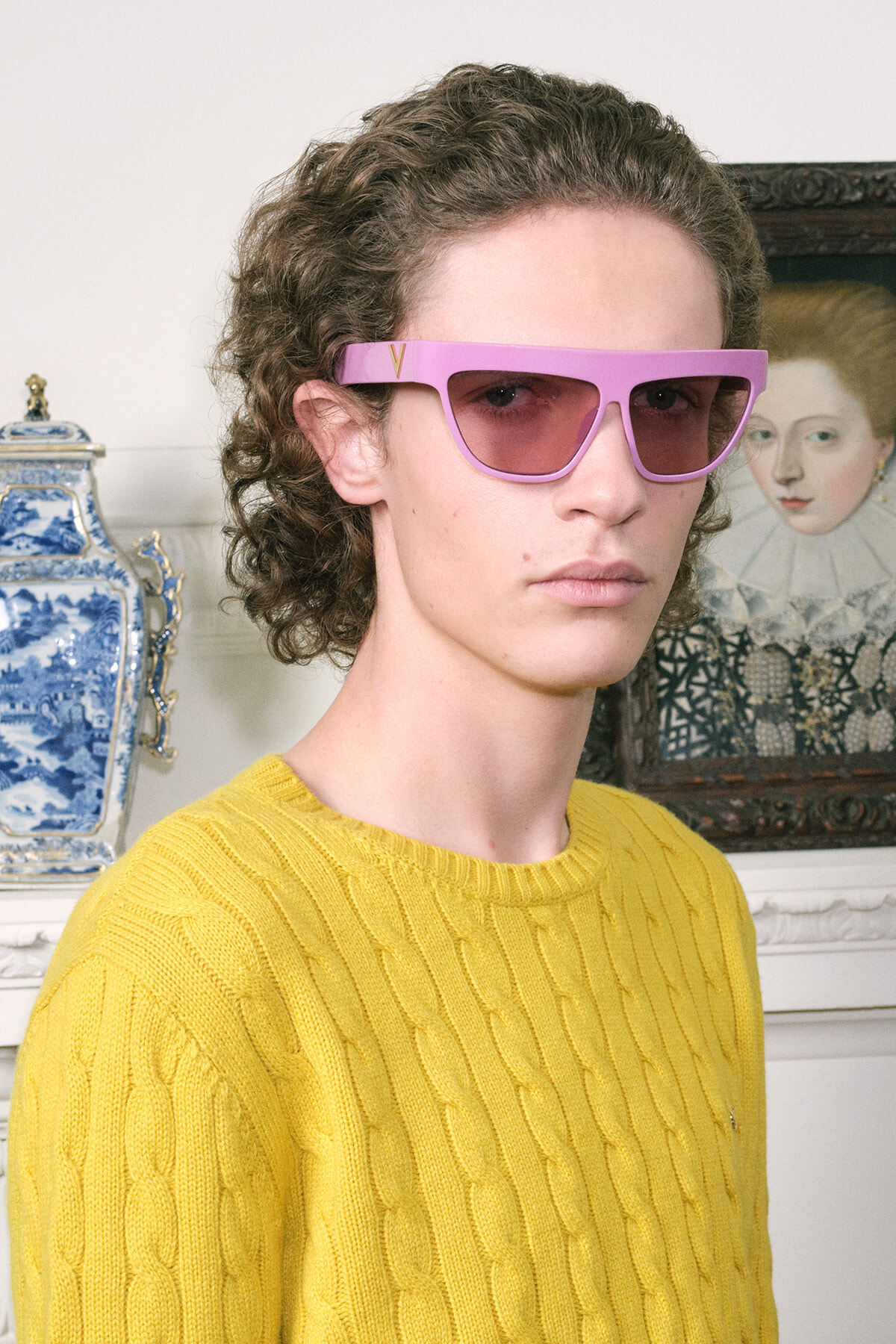 Person with short curly hair wearing a bright yellow cable-knit sweater and pink rectangular sunglasses indoors, with blue-and-white porcelain vase in the background.