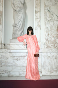 Woman in a pink floor-length dress with puff sleeves, black sunglasses, and a brown clutch, posing beside a marble statue.