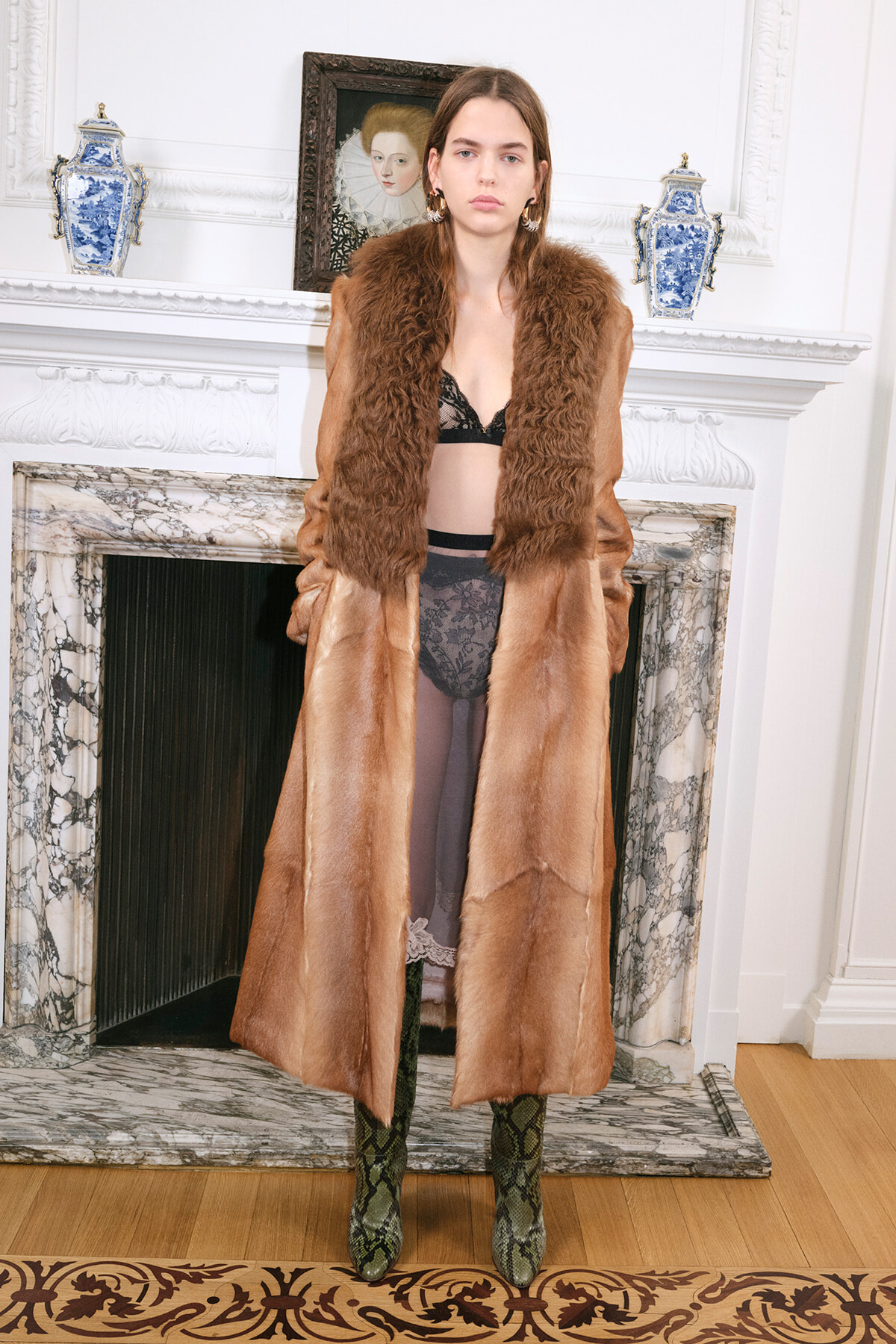 Woman wearing a long brown fur coat over black lace lingerie, standing in a marble fireplace room with blue and white vases on the mantel.