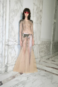Model in a blush beaded evening gown with silver embroidery and a satin belt, standing in a marble hallway wearing pink sunglasses.