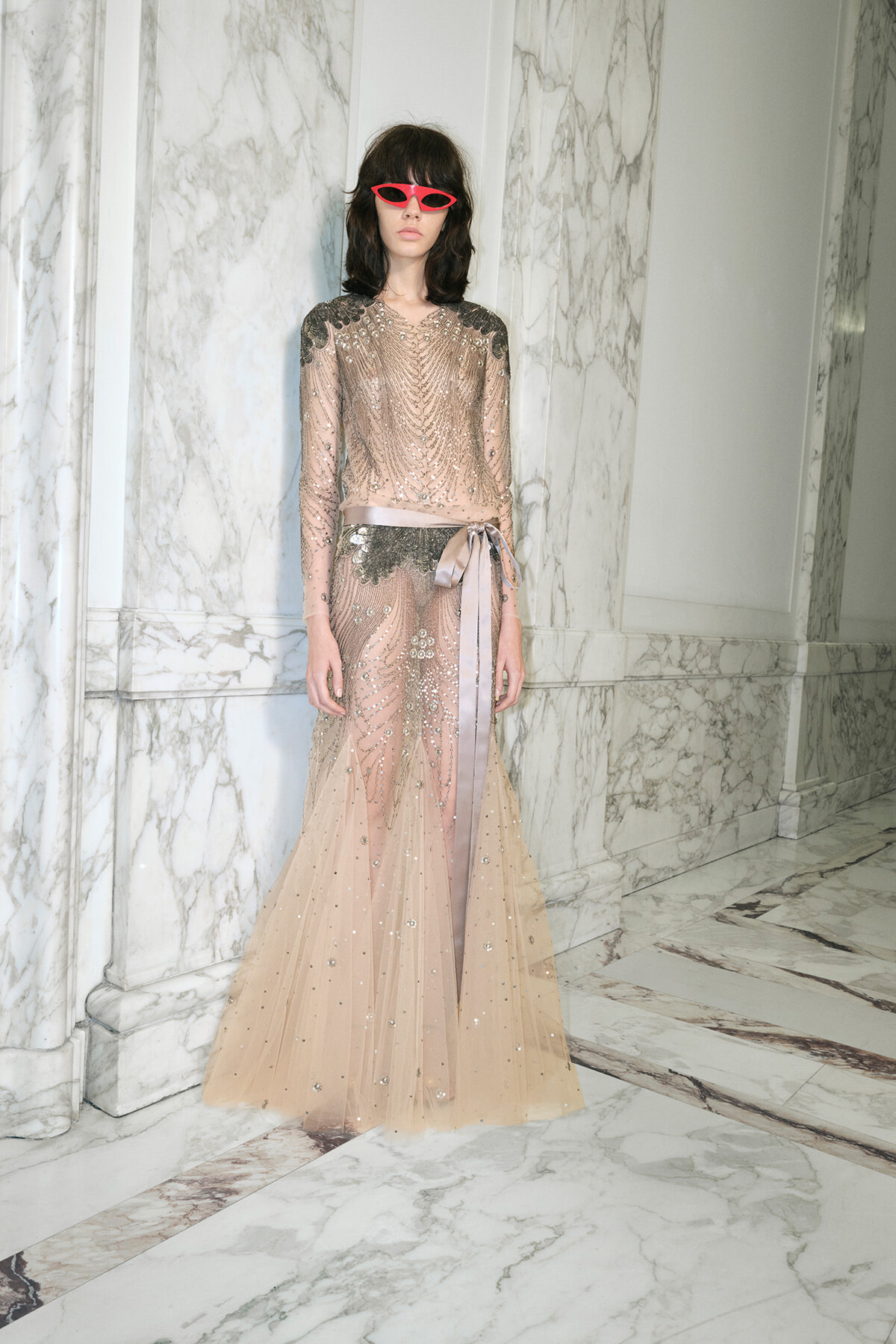 Model in a blush beaded evening gown with silver embroidery and a satin belt, standing in a marble hallway wearing pink sunglasses.
