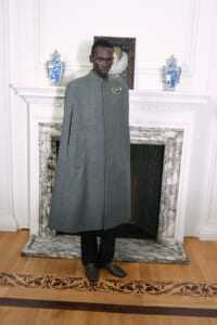 Man wearing a knee-length gray coat with a high collar and a decorative brooch, standing in a room with a marble fireplace and blue-white vases behind him.
