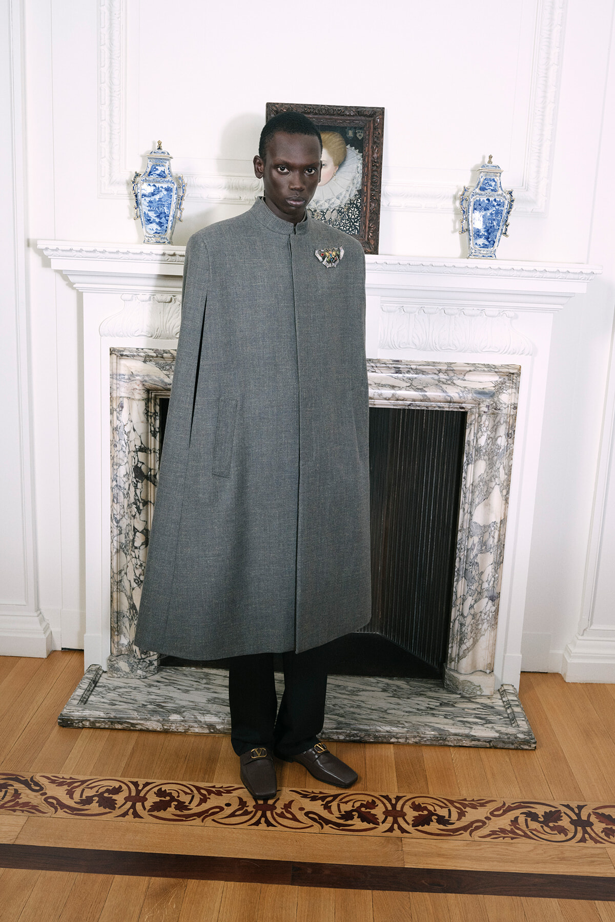 Man wearing a knee-length gray coat with a high collar and a decorative brooch, standing in a room with a marble fireplace and blue-white vases behind him.