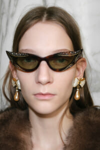 Woman wearing large cat-eye sunglasses with rhinestone accents and a fur collar; gold bow earrings with pearl drops.
