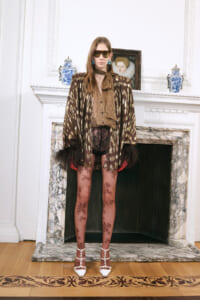 Model wearing a brown-gold animal-print cape over a leopard blouse, sheer floral tights, white studded heels, and oversized sunglasses by a marble fireplace.