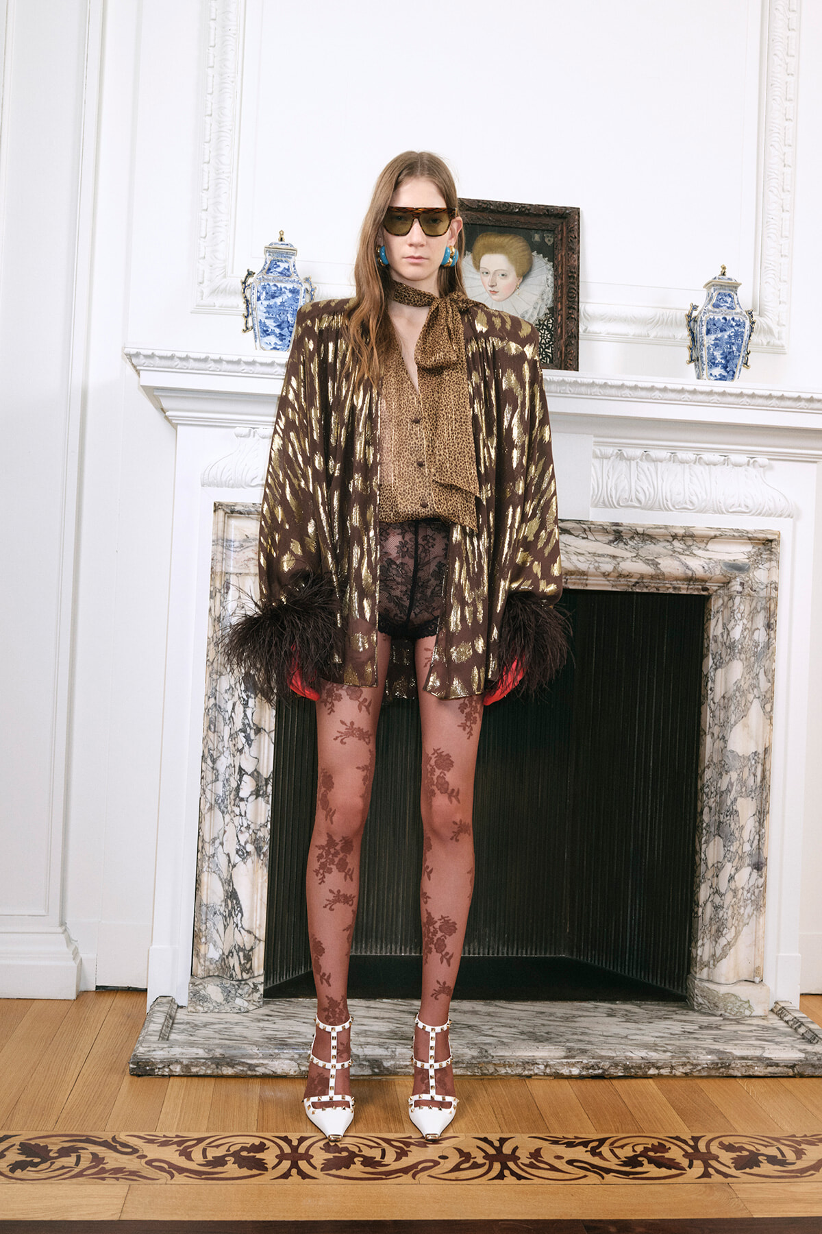 Model wearing a brown-gold animal-print cape over a leopard blouse, sheer floral tights, white studded heels, and oversized sunglasses by a marble fireplace.