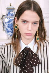 Fashion portrait of a young woman wearing oversized gold earrings with green stones, a white collared shirt, and a brown polka-dot bow over a striped blouse, with a blue-and-white vase in the background.