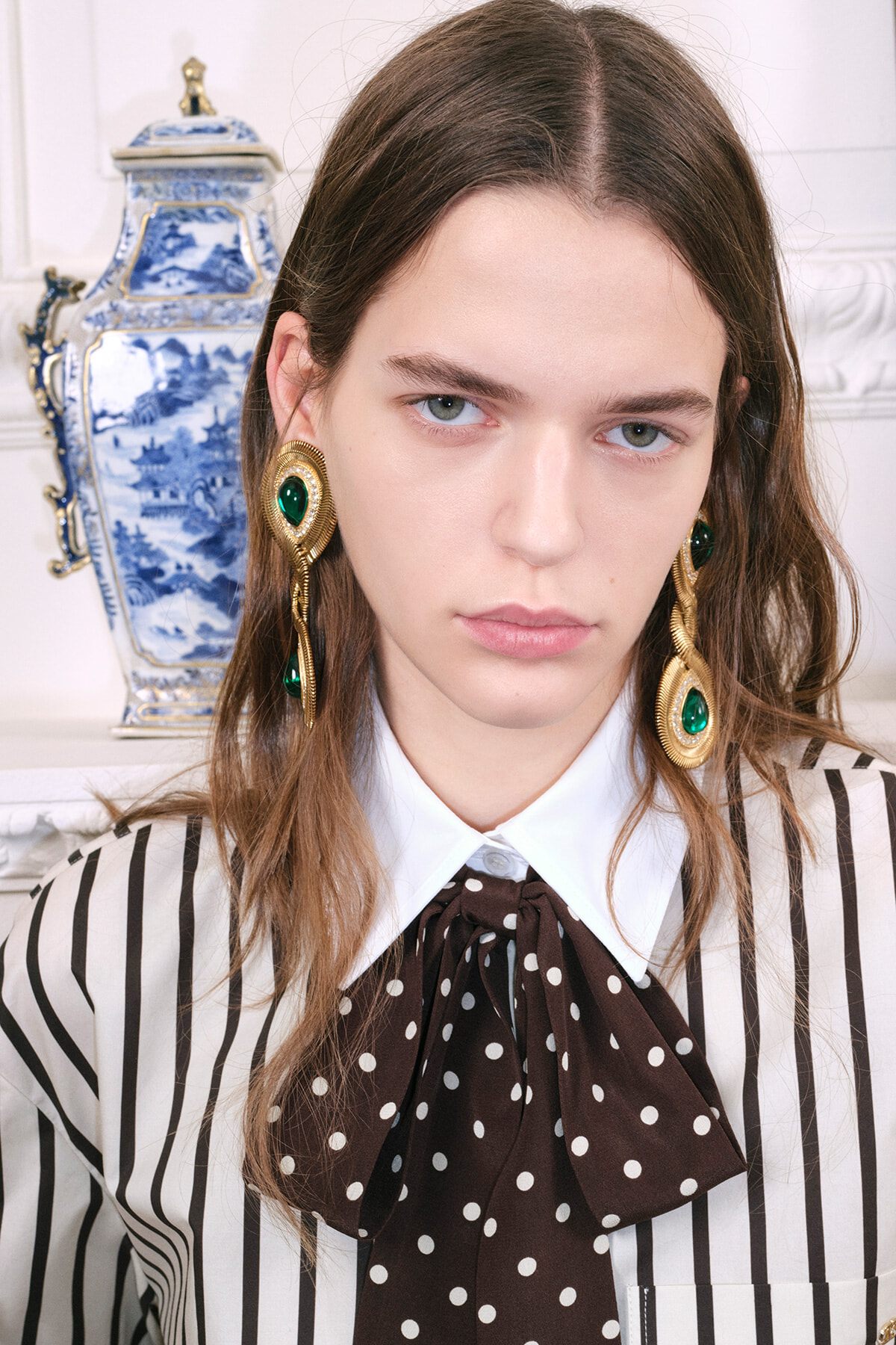 Fashion portrait of a young woman wearing oversized gold earrings with green stones, a white collared shirt, and a brown polka-dot bow over a striped blouse, with a blue-and-white vase in the background.