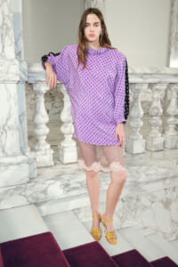Model in a lavender oversized polka-dot tunic with black side stripe, sheer lace-trim shorts, and yellow strappy heels posing on a marble staircase.