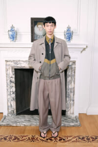 Full-body portrait of a person standing in a beige overcoat over a color-block zip-up jacket and wide taupe trousers, in a room with marble fireplace and decorative vases on the mantel.