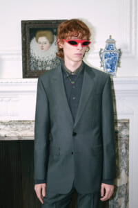 Young man in a dark green suit and red sunglasses stands in a refined room with a porcelain blue vase on a mantel behind him.