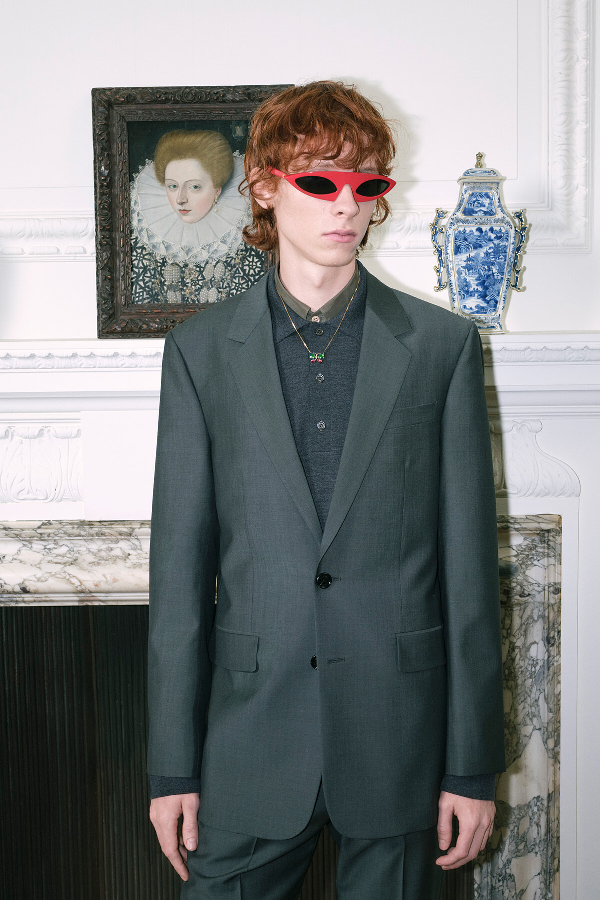 Young man in a dark green suit and red sunglasses stands in a refined room with a porcelain blue vase on a mantel behind him.