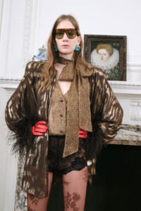 Model wearing a metallic brown coat with a leopard scarf, red gloves, and oversized sunglasses indoors with a framed portrait in the background?