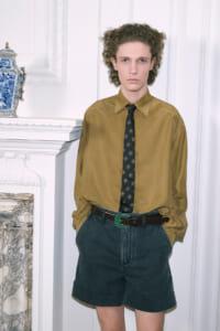 Young male model in a mustard shirt, patterned tie, and dark shorts posing in a white ornate room.