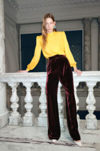 Model wearing a yellow satin blouse with puff sleeves and burgundy velvet wide-leg pants, leaning on a marble railing in an ornate interior of a grand hall.