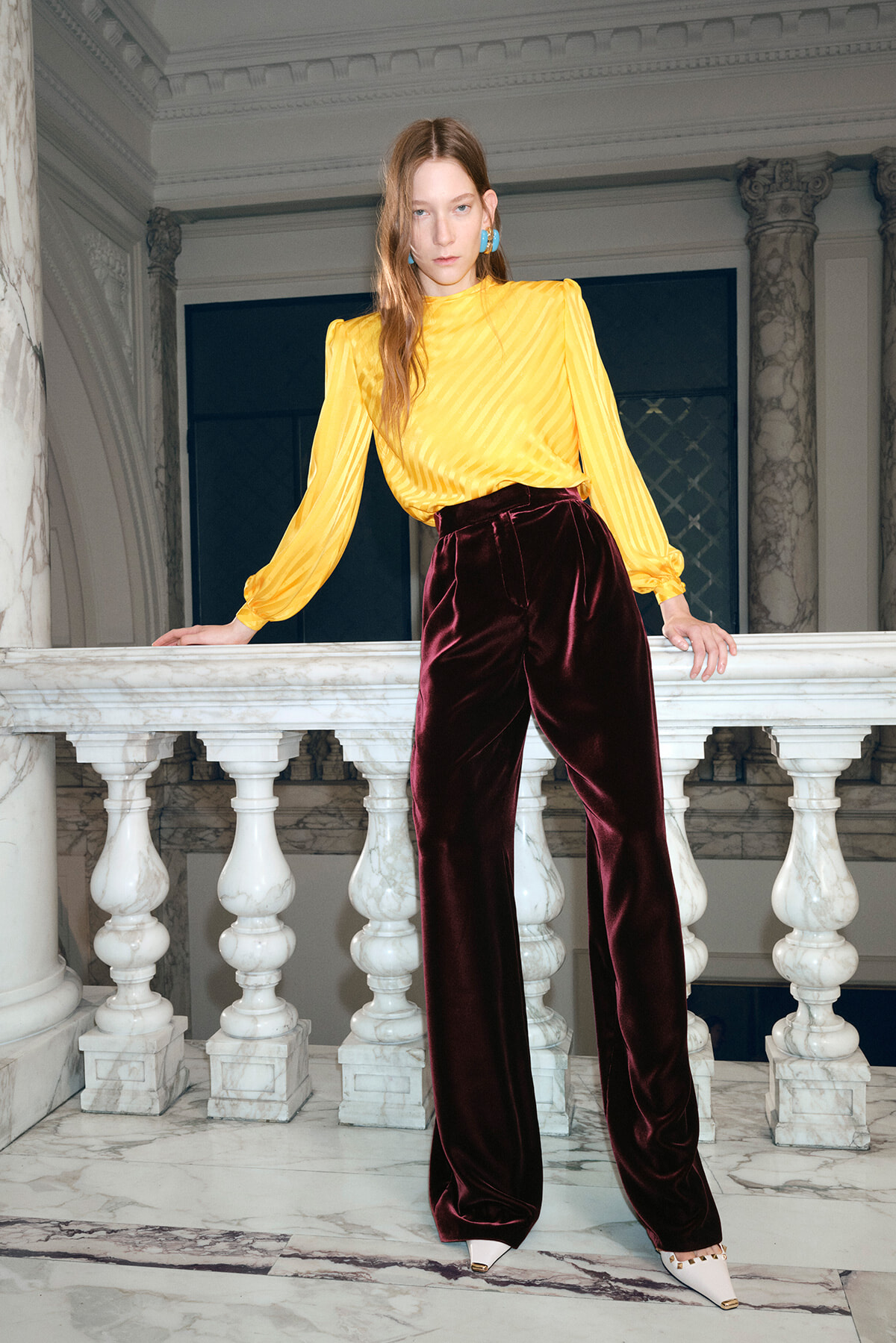 Model wearing a yellow satin blouse with puff sleeves and burgundy velvet wide-leg pants, leaning on a marble railing in an ornate interior of a grand hall.