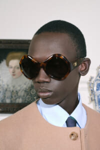 Person wearing oversized tortoiseshell sunglasses, light-blue shirt, and tan coat with a buttoned lapel.