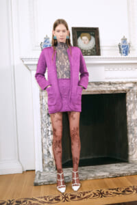 Model wearing a purple quilted coat over a sheer lace top, standing in a refined room with a marble fireplace and decorative vases in the background.”