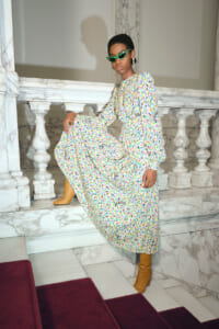 Woman in a long multicolor printed dress and tan boots posing on a marble staircase with green cat-eye sunglasses.