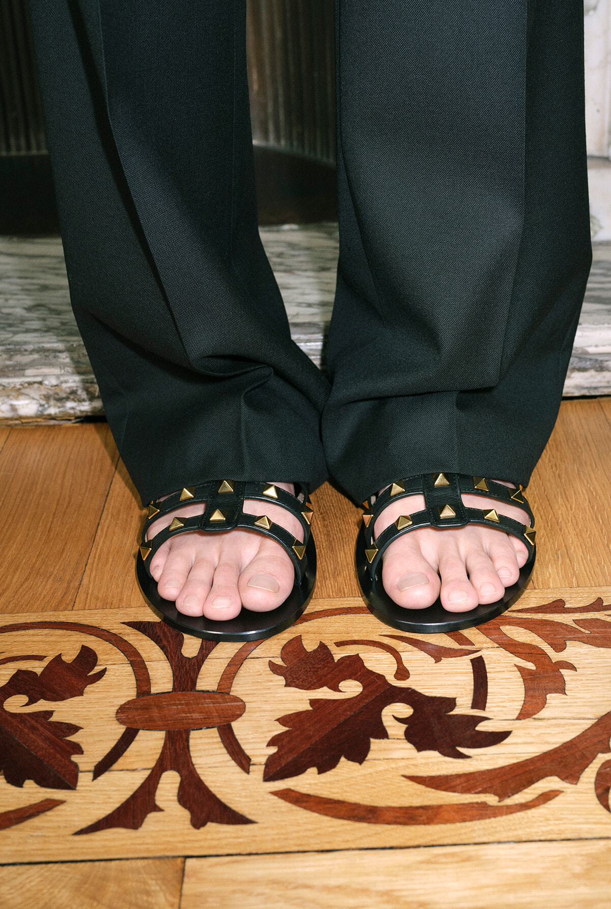 Feet in black studded sandals standing on a wooden floor with an ornate pattern showing toes clearly visible.