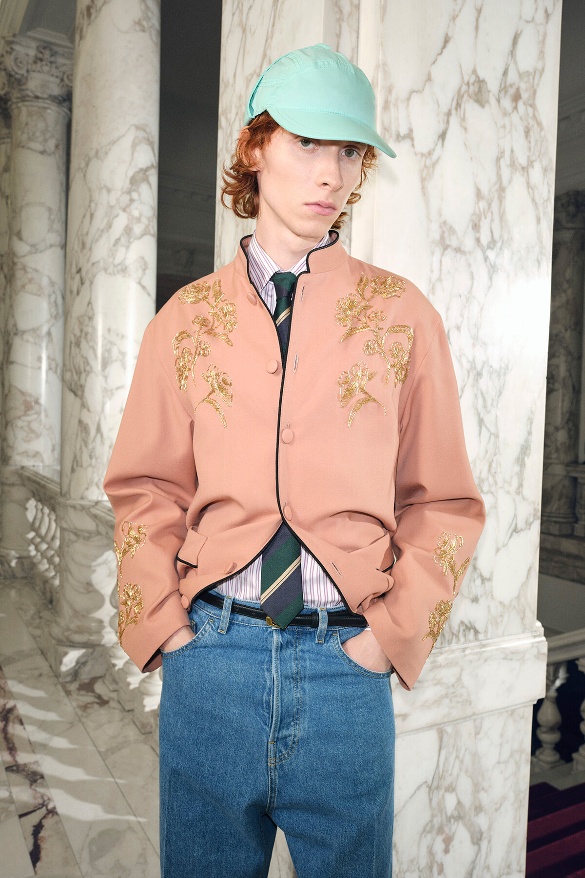 Young man wearing a peach embroidered jacket, striped shirt, green tie, and jeans, with a mint cap, posing in a marble hall.